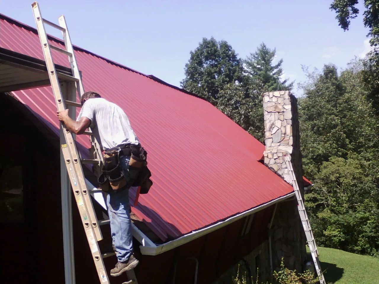 Roofer on a ladder beside a red metal roof and stone chimney — Soffit & Fascia Repair by Roofing Brothers in Lumberton